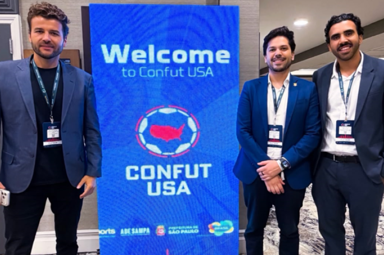 PBH participa de encontro estratégico sobre a Copa do Mundo Feminina de 2027 Três homens vestindo ternos azuis, sendo um deles com camisa preta e os outros dois com camisas sociais brancas, posando ao lado de um cartaz com fundo azul com os seguintes dizeres: Welcome Confunt USA.