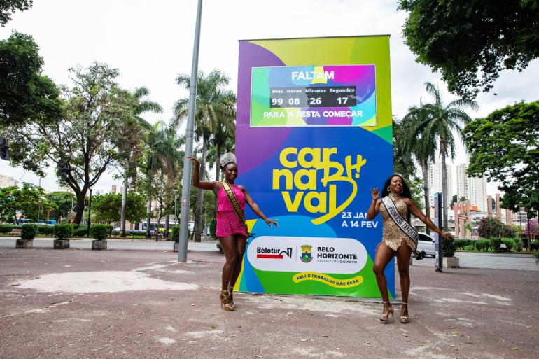 Totem marca contagem regressiva de 100 dias para o Carnaval de BH 2027 Totem colorido do Carnaval de Belo Horizonte, nas cores amarelo, verde, roxo e azul, com a marca “Carnaval de BH” e contador eletrônico que marca a contagem regressiva de 100 dias para a folia. Em frente ao totem duas mulheres, caracterizadas com roupa carnavalesca em posição de dança.