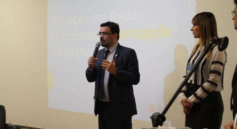 O controlador-geral do município, Leonardo Ferraz, palestra em evento, em frente a telão, ao lado de mulher. 