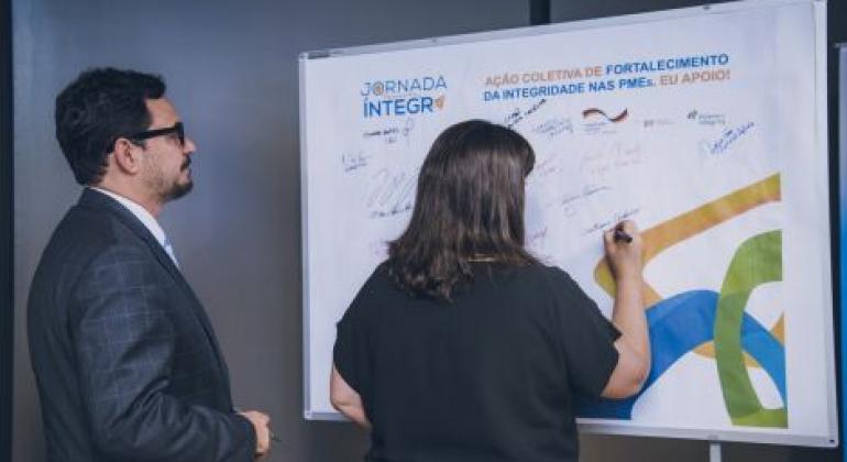 Projeto que estimula práticas de integridade em pequenas e médias empresas é apoiado pela Prefeitura de Belo Horizonte. Foto: Divulgação PBH Um homem em uma mulher assinam em painel da Jornada Íntegra: um bom negócio para a sua empresa!”.