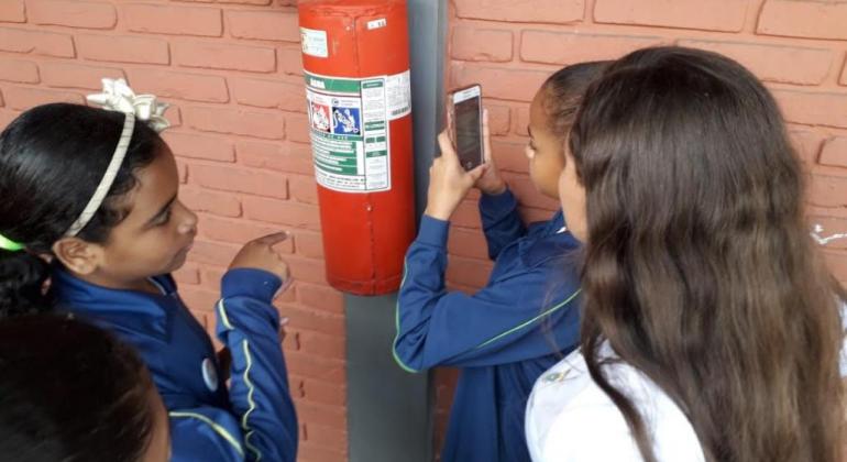 Quatro estudantes fiscalizam extintor de incêndio em escola municipal, durante o dia. 