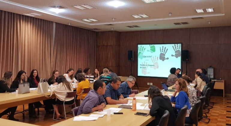 Durante o evento, práticas e ações de capacitação teóricas sobre o tema foram promovidas por Hevellyn Albres, do Ministério do Trabalho. Foto: Divulgação PBH Mais de 18 pessoas em mesa em forma de "u", com projeçao à frente.