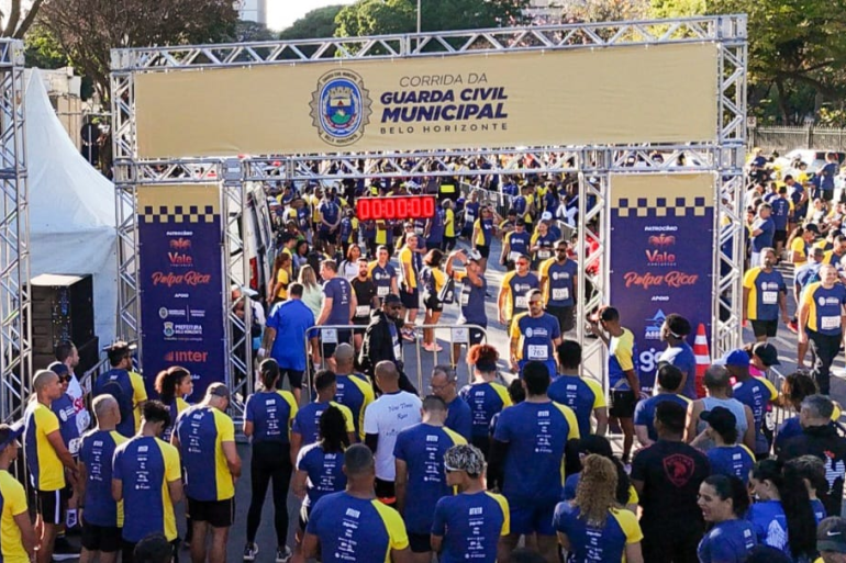 Multidão em frente a uma estrutura de metal com banner da Guarda Municipal se preparando para iniciar a corrida 