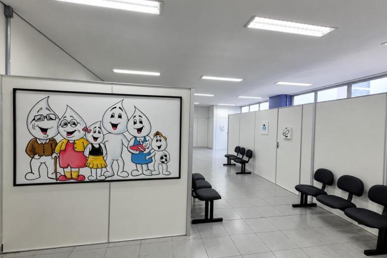 Corredor de unidade de saúde com cadeiras pretas com mural de vacinação