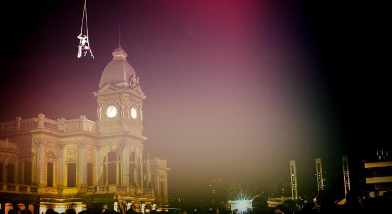 Multidão assiste a show da Virada Cultural na Praça da Estação, durante a noite; mulher realiza performance com cabos, "pendurada no ar", sentada em banco. 