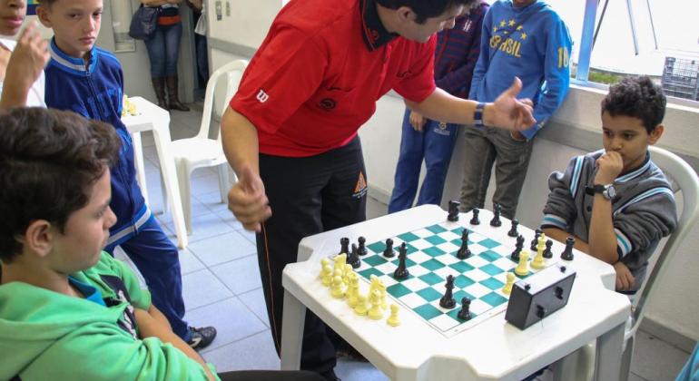 Crianças jogam xadrez acompanhada por um adulto.