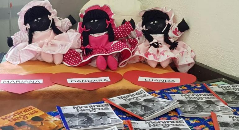 Três bonecas pretas com touca e vestido e identificadas com nomes diferentes - Mariana, Dandara e Luanda - sobre uma mesa. À frente, o livro "Meninas negras" com várias cópias. 