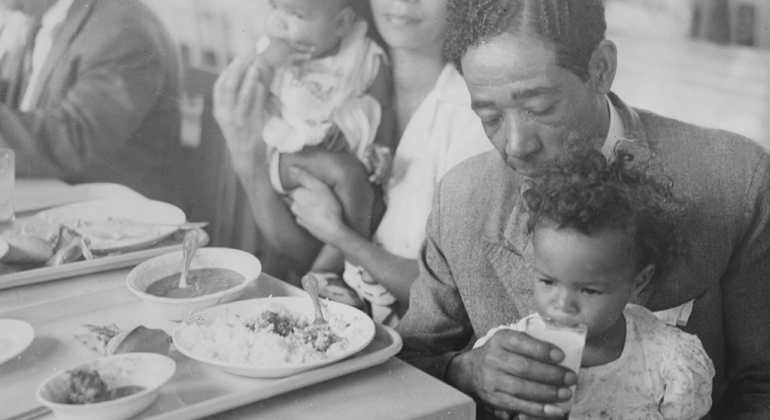 Em foto P&B de acervo do Museu Histórico Abílio Barreto, homem em frente à mesa alimenta criança que está sentada em seu colo.