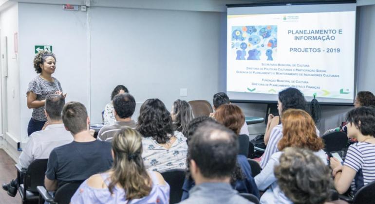Servidores e gestores da Secretaria e da Fundação Municipais de Cultura se reúnem em capacitação para otimizar ações em 2019. Foto: Ricardo Laf/PBH Mais de catorze pessoas, sentadas, assistem a exposição de mulher na capacitação “Planejamento e Informação – Projetos 2019”.