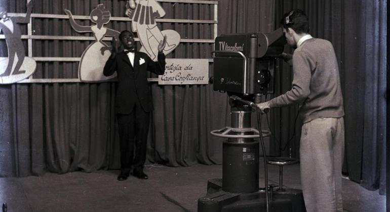 Foto em preto e branco feita em um estúdio de televisão. Um homem está em frente a uma câmera enquanto outro manuseia o equipamento