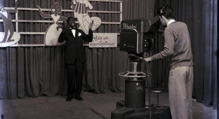 Museu da Imagem e do Som revive a história da TV Itacolomi em nova exposição Foto em preto e branco feita em um estúdio de televisão. Um homem está em frente a uma câmera enquanto outro manuseia o equipamento