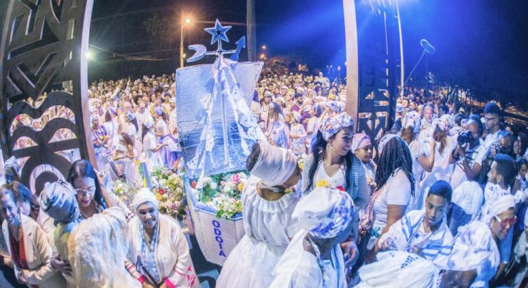 Mais de 300 pessoas, a maioria de branco e azul com turbante na cabeça e com um estardarte de Iemanjá, confraternizam na orla da Lagoa da Pampulha, à noite. 