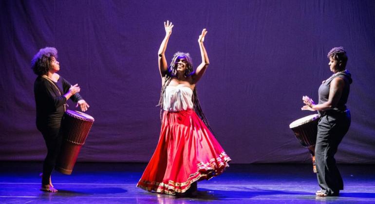 Mulher dança no palco enquanto duas outras tocam instrumento de percussão