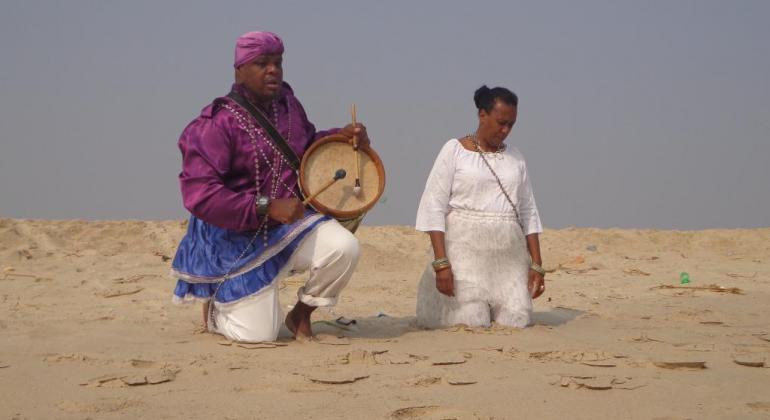 Homem negro ajoelhado toca tambor ao lado de mulher negra com roupa branca ajoelhada, na areia, durante o dia. 