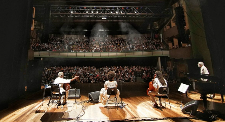 O Cine Theatro Brasil Vallourec sediou a abertura oficial da 10ª edição do FAN-BH, na segunda, dia 18/11. TReês artistas negros se apresentam no palco; ao fundo, uma platéia lotada assiste.
