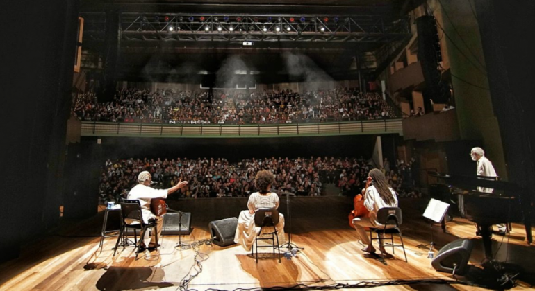 TReês artistas negros se apresentam no palco; ao fundo, uma platéia lotada assiste. 