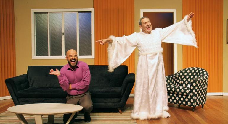 Homem vestido de anjo, de pé, ao lado de homem sentado no sofá, em cenário de peça teatral. 