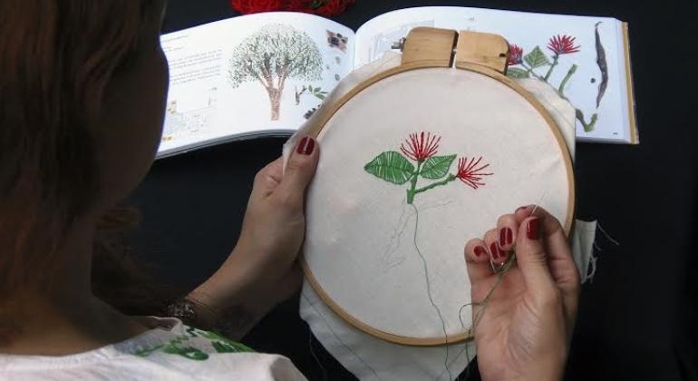 Bordados produzidos nas oficinas do projeto “Bordando Memórias” são expostos no Museu Casa Kubitschek. Foto: Divulgação PBH Mão feminina borda em aro circular flor de um livro ilustrado.