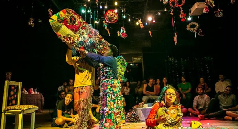 Espetáculo “Eles também falam de amor” está em cartaz no teatro Marília de 8 a 10 de novembro e tem entrada gratuita. Foto: Netun Lima três atores interagem em cenário colorido, com cadeira, cama e papéis coloridos pendurados no teto.