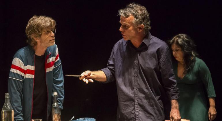 O espetáculo “Nós” está em cartaz de 30/5 a 2/6 ; “Outros”, de 5 a 9/6. Às sextas, as sessões terão interpretações em libras. Foto: Guto Muniz Homem mostra faca para outro homem; mulher com vestido está atrás, à direita.