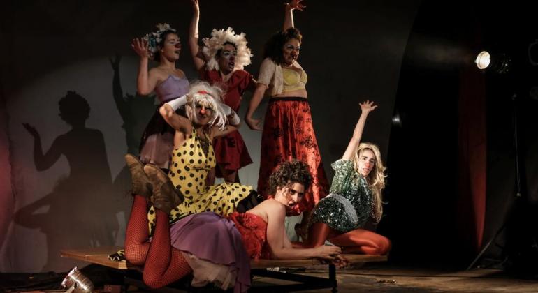 Seis mulheres vestidas de palhaças posam em apresentação teatral. 