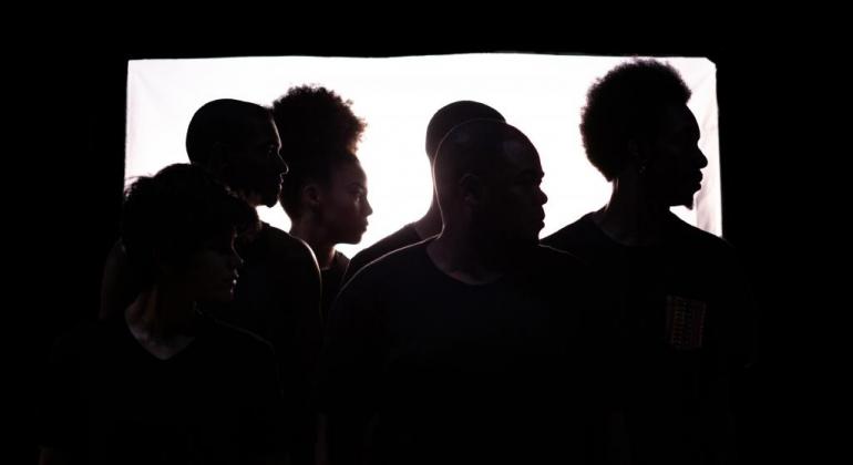 A apresentação de dança utiliza a linguagem das danças urbanas, elemento da cultura hip hop, para tematizar a vida nas favelas brasileiras. Foto: Pablo Bernardo Quatro perfis humanos da contraluz, formando perfis negros em um quadrado negro.