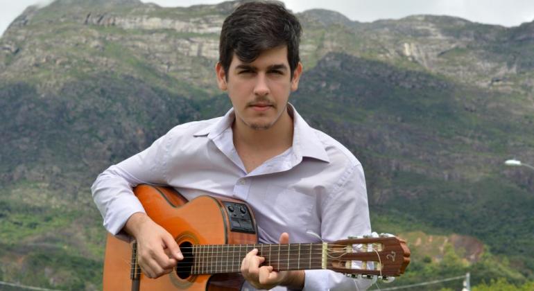 Homem de camisa com violão, tendo ao fundo montanha, a céu aberto, durante o dia. 