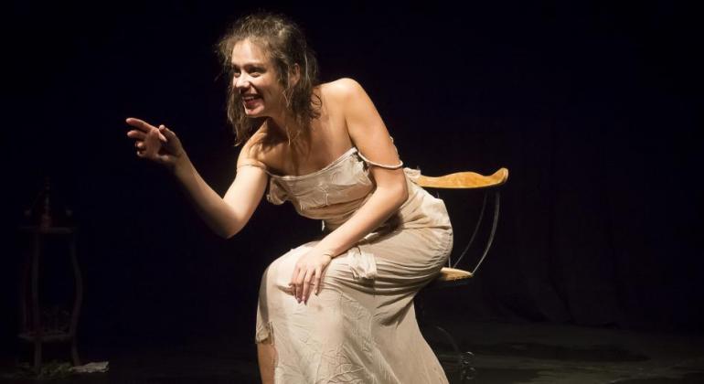 O Teatro Marília será palco, de 10/5 a 2/6, da segunda edição da mostra “Nos Porões da Loucura”. Foto: Guto Muniz Mulher de vestido com as alças caídas, sentada em cadeira no palco, fala.
