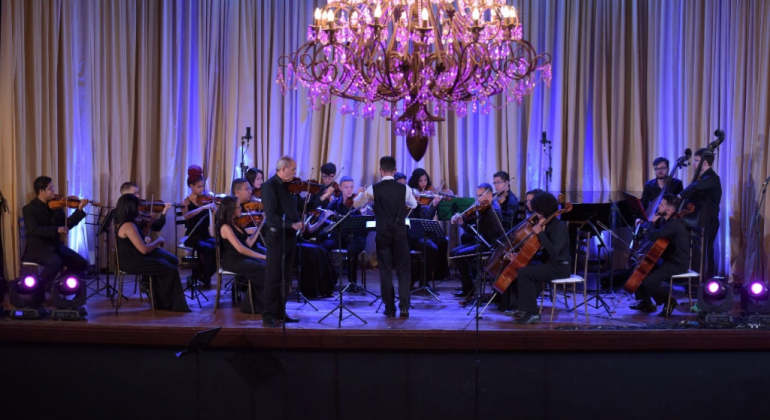 Mais de doze músicos da Orquestra de Cordas se apresentam em palco com lustre de cristal imponente pendurado, à meia luz. 