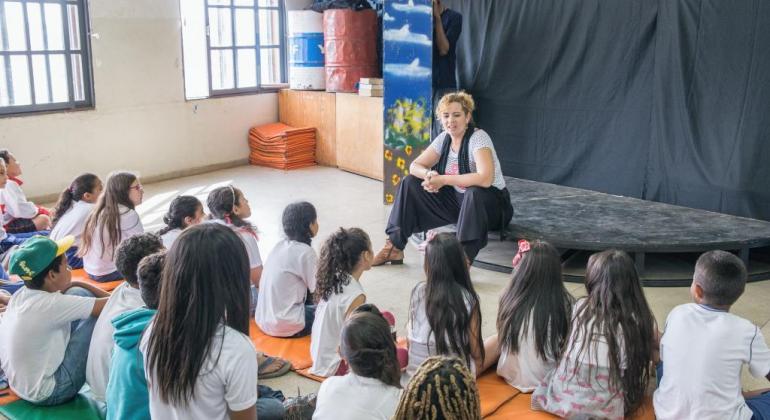 Mulher, sentada à frente de um palco palestra para mais de quinze crianças, também sentadas. 