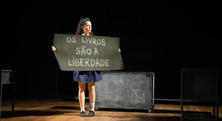 O espetáculo gratuito “Classe”, do Grupo Mulheres Míticas, está em cartaz no Teatro Raul Belém Machado nos dias 9 e 10/11. Foto: Raquel Carneiro Mulher de saia azul, ao lado de quadro negro encostado no chão, com uma placa com os seguintes dizeres: "Os livros são a liberdade".