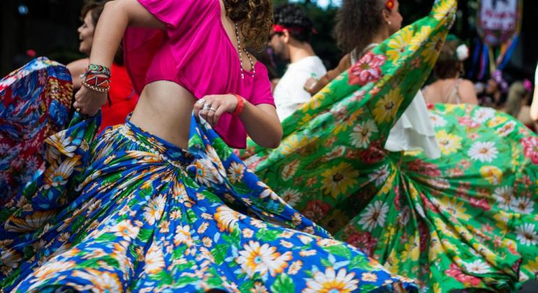 Duas mulheres dançam e agitam saias coloridas de chita, durante o dia. 