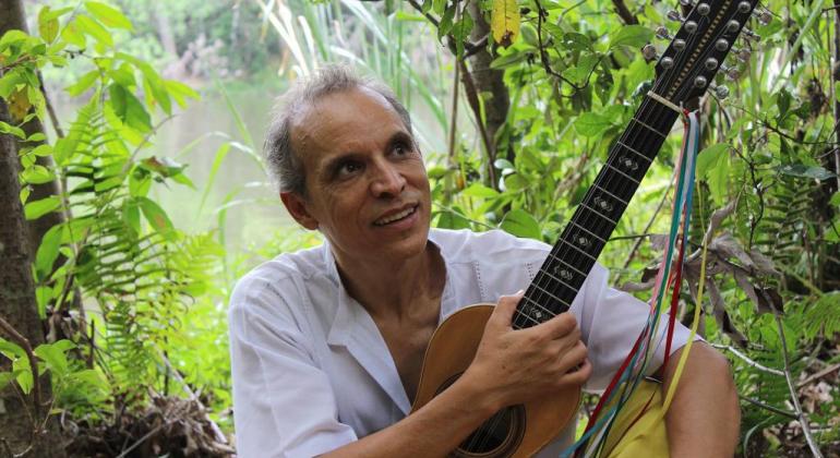 Homem sentado com viola nos braços; ao fundo, muitas plantas e árvores. 