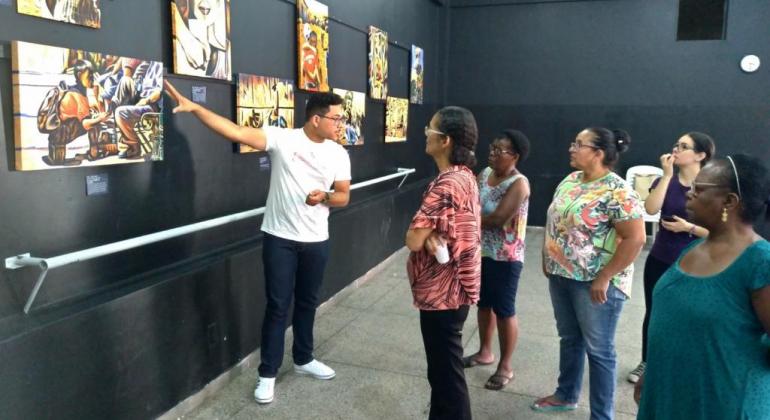 Homem aponta para um dos sete quadros expostos na parede, acompanhado de cinco pessoas, em uma sala. 