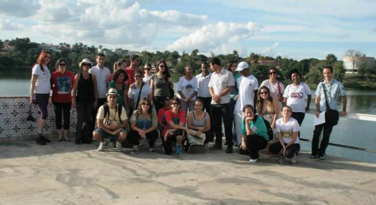 Mais de vinte pessoas , a maioria de pé e a minoria sentada à frente, posam para foto tendo a lagoa da Pampulha ao fundo. 