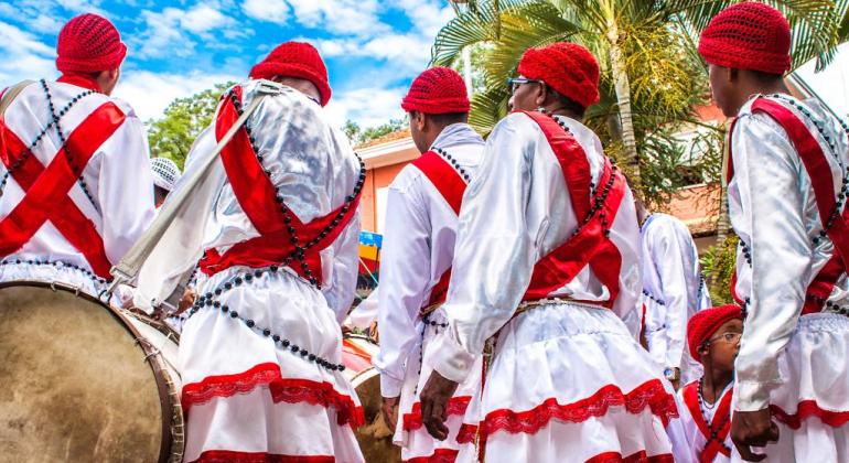 Cinco musicistas de cultura popular, de costas, com vestes vermelha e preta e turbante vermelho, um deles está com um tambor. 