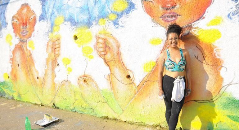 Até o dia 15/9, a Lagoinha é palco para mais uma edição do Circuito Urbano de Arte, que está levando à região as intervenções artísticas. Foto: Divulgação PBH Mulher posa à frente de muro grafitado, durante o dia.