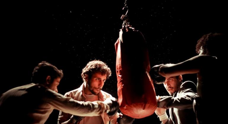 O Teatro Francisco Nunes recebe, entre os dias 7 e 9/11, às 20h, o espetáculo “É só uma formalidade”, do Grupo Grupo Quatroloscinco. Foto: Núbia Abe Três homens revezam socos em saco de lutador de boxe em palco, à meia luz.