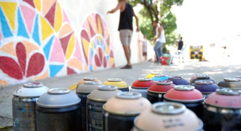 Latas de spray em primeiro plano e, ao fundo, um homem que pinta um muro.