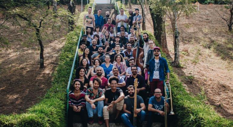 Mais de quarenta músicos da Geraes Big Band sentados em uma escada de um local aberto e com verde ao redor, durante o dia. 