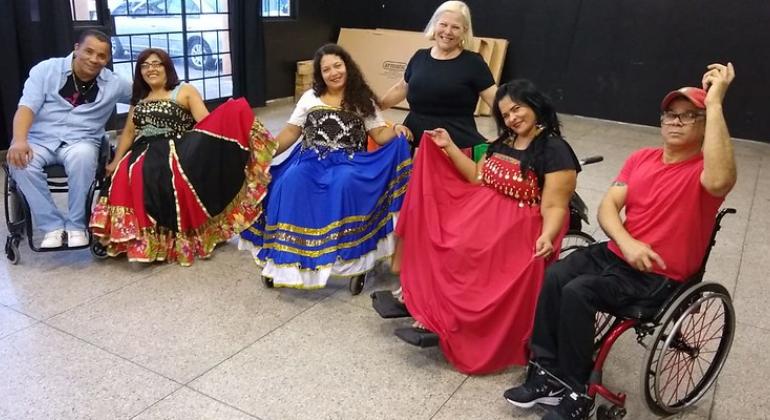 O Centro Cultural Pampulha iniciou em novembro a oficina “Movimento e Sensibilização”, para proporcionar aos cadeirantes a formação em dança. Foto: Divulgação PBH Cinco cadeirantes, dos quais três são mulheres com vestidos bordados, e dois homens, acompanhados de uma mulher, de pé.