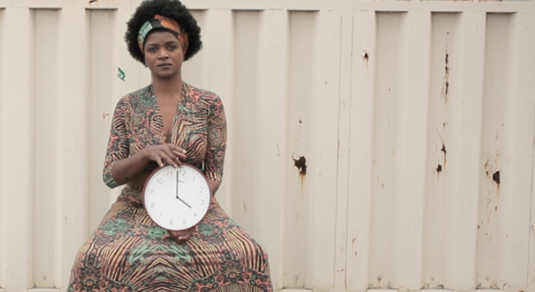 Foto do trabalho de Dayane tropicaos, com mulher negra de vestido, sentada em frente à portão, com um relógio na mão. 
