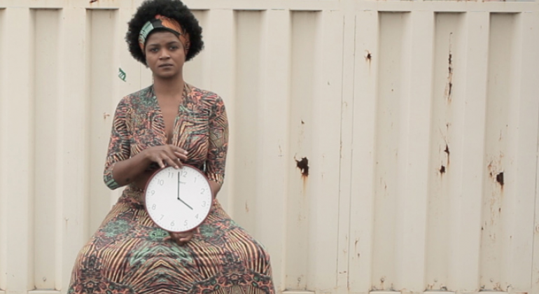 A Prefeitura de Belo Horizonte inaugura no dia 14/9, sábado, às 14h, no Museu de Arte da Pampulha, a exposição 7º Bolsa Pampulha. Foto: Divulgação PBH Foto do trabalho de Dayane tropicaos, com mulher negra de vestido, sentada em frente à portão, com um relógio na mão.