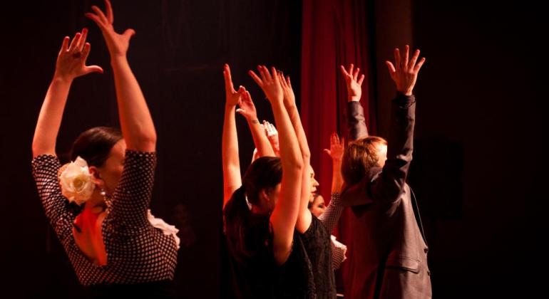 Duas mulheres e dois homens erguem as mãos, em um gesto tipico da dança flamenca, sob luz avermelhada. 