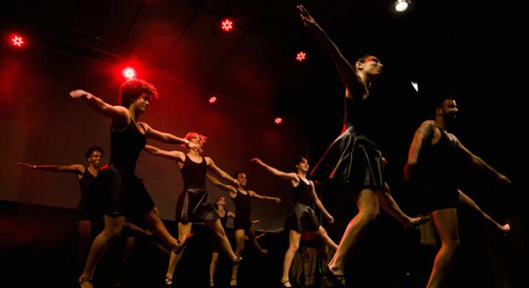 Sete bailarinos, quatro mulheres com saia e camiseta pretas, e os três homens com com short e camiseta pretos, dançam, de maneira coordenada, em palco teatral com luz vermelha..