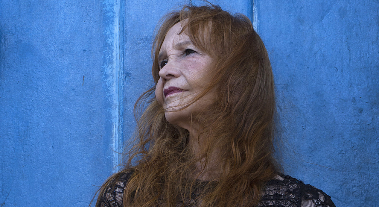 Foto da cineasta Helena Ignez sobre parede de fundo azul. 