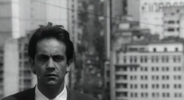 Imagem em P&B do ator Paulo José, muito jovem, de terno, com prédios ao fundo. Cena do filme A Vida Provisória.