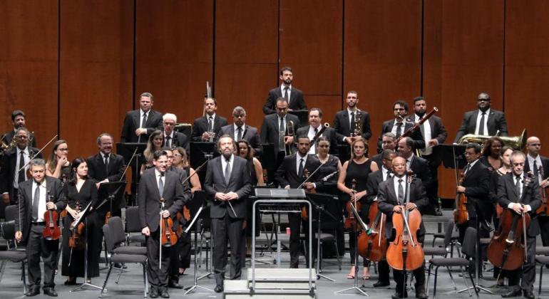 Imagem de todos os componentes da Orquestra Sinfônica no palco de um teatro