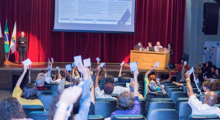 mais de trinta pessoas de mãos levantadas, votando, no Teatro Marília; ao fundo, mesa de abertura de evento e telão.