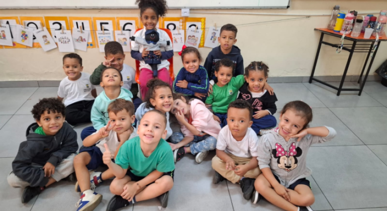 #paratodosverem: a foto mostra crianças em uma sala de aula. Com exceção de uma menina, a turma está sentada no chão. A aluna, ao centro, senta-se em uma cadeira e segura o boneco Tobias, que tem pele negra e usa blusa azul clara, shorts, calçados e uma boina azul escura. Ao fundo, há um quadro branco e cartazes com palavras acompanhadas de uma ilustração.